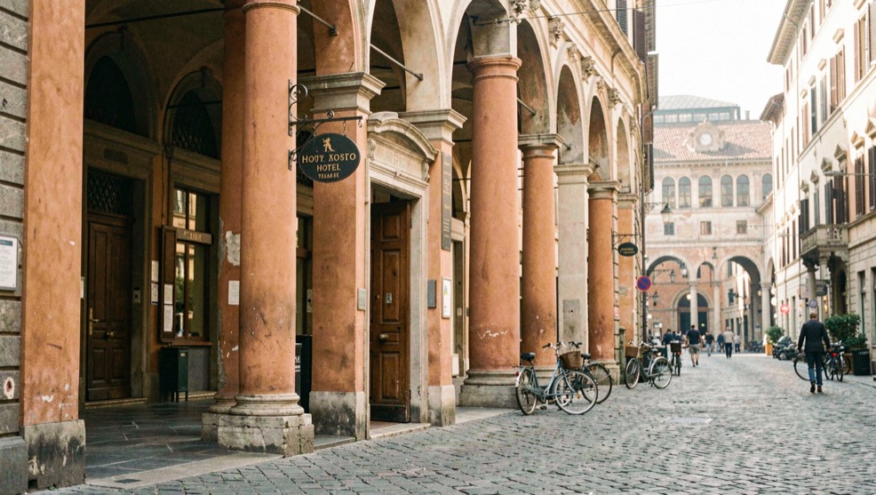 Porticoed Bologna street in soft morning light with ochre columns, a small hotel sign above a wooden door and bicycles passing toward the station