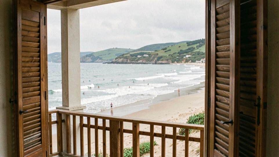 Sea front pension balcony at Zarautz with wooden shutters half open onto a long bay beach, surfers below and green hills closing the horizon